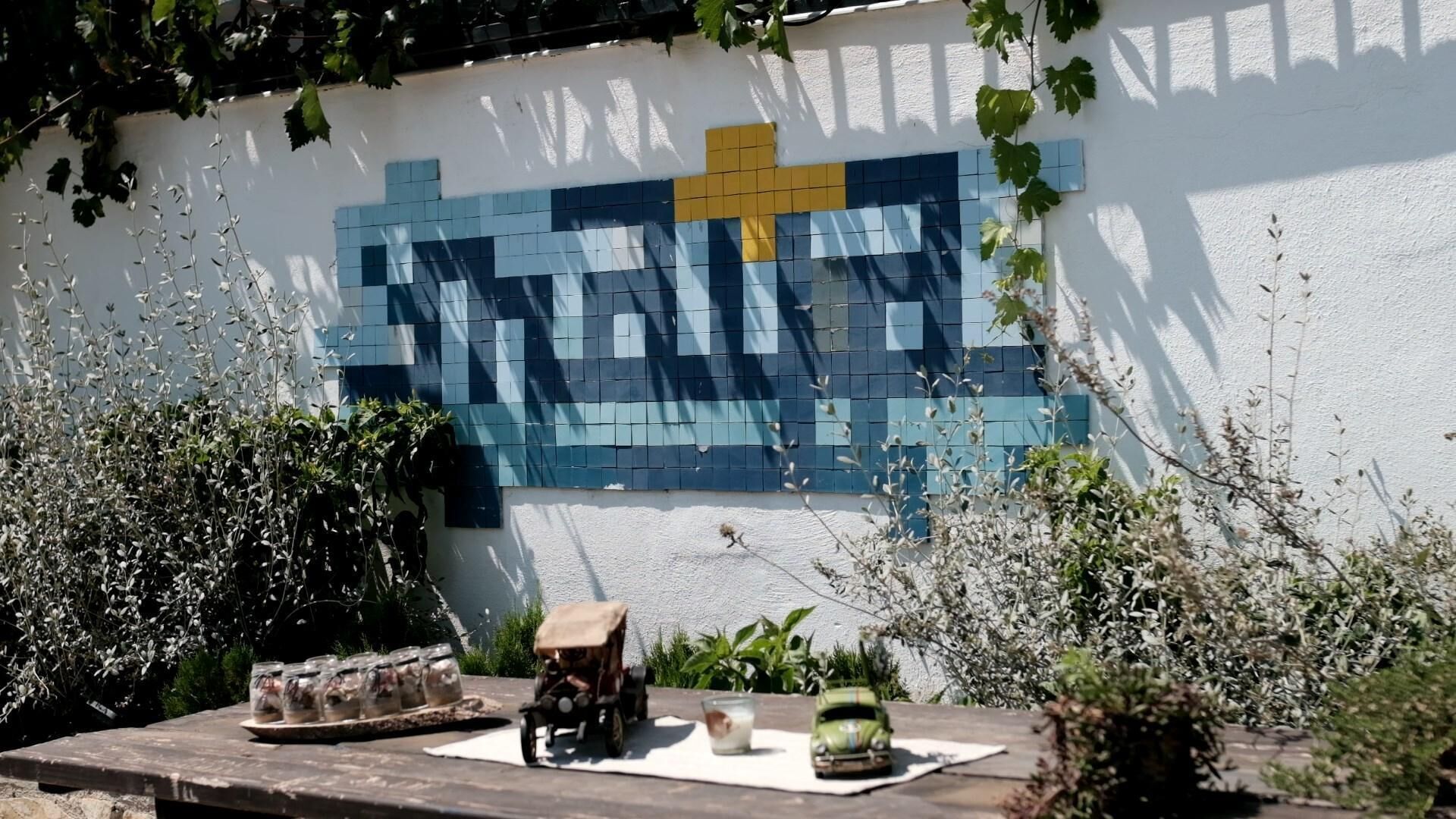 A wall with a mosaic pattern, a wooden table with a model car and a glass, and surrounding greenery.