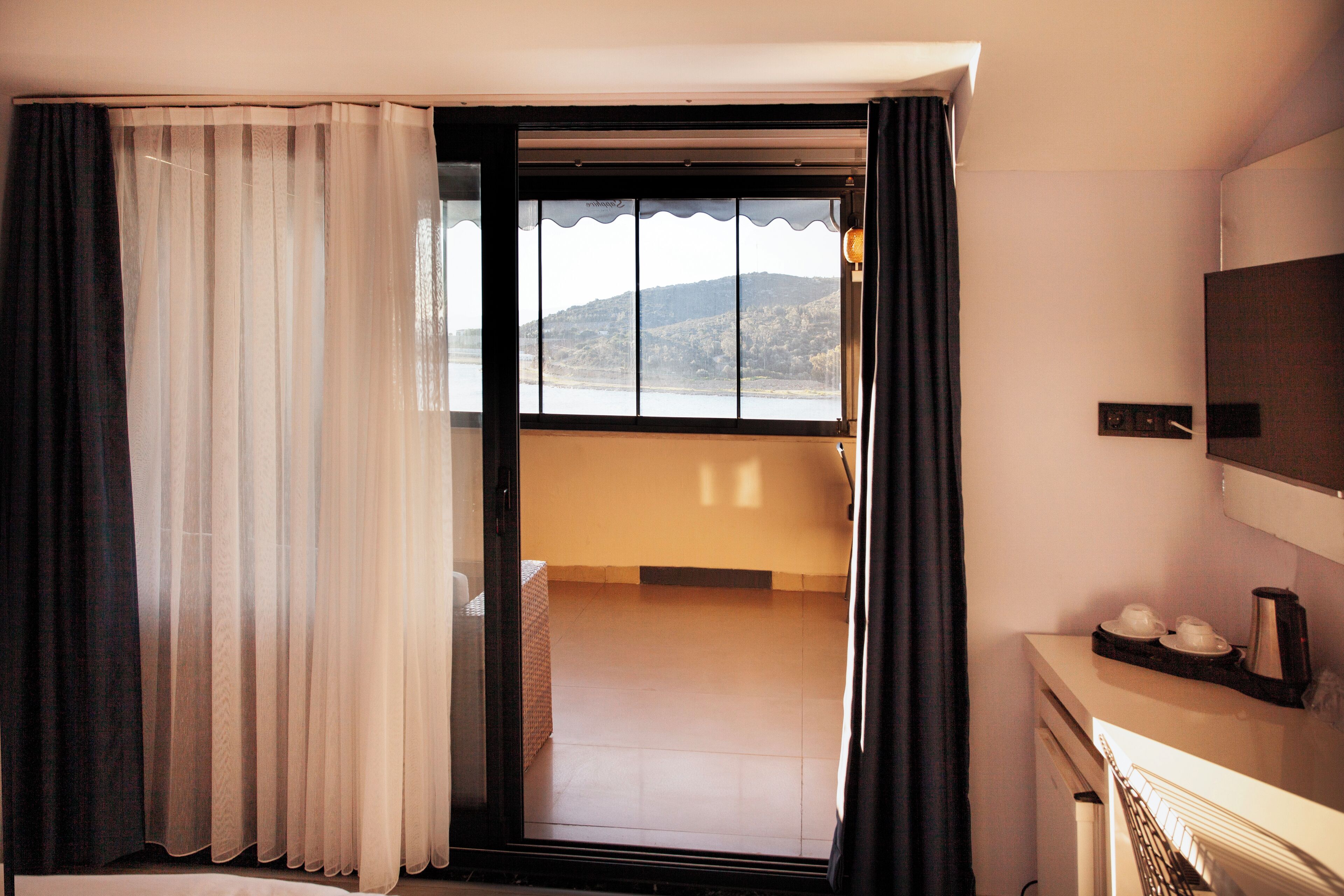 A hotel room with a sliding glass door leading to a balcony with a view of hills. There is a small table with cups and a kettle on it.