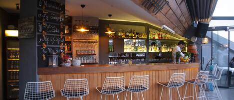 A bar area with wooden bar counter, white wire mesh bar stools, and a variety of bottles and glasses on shelves.