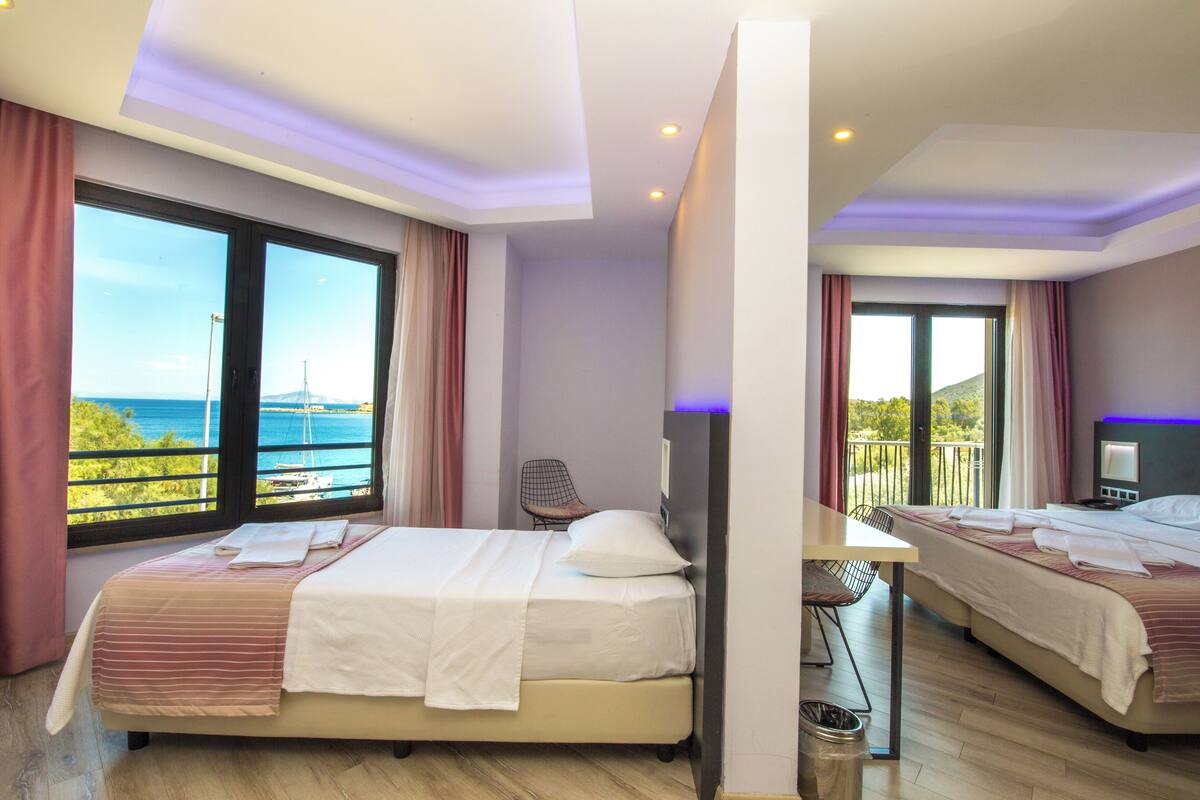 A hotel room with two beds, a desk, and a chair. The room has large windows with a view of the sea and a bridge.