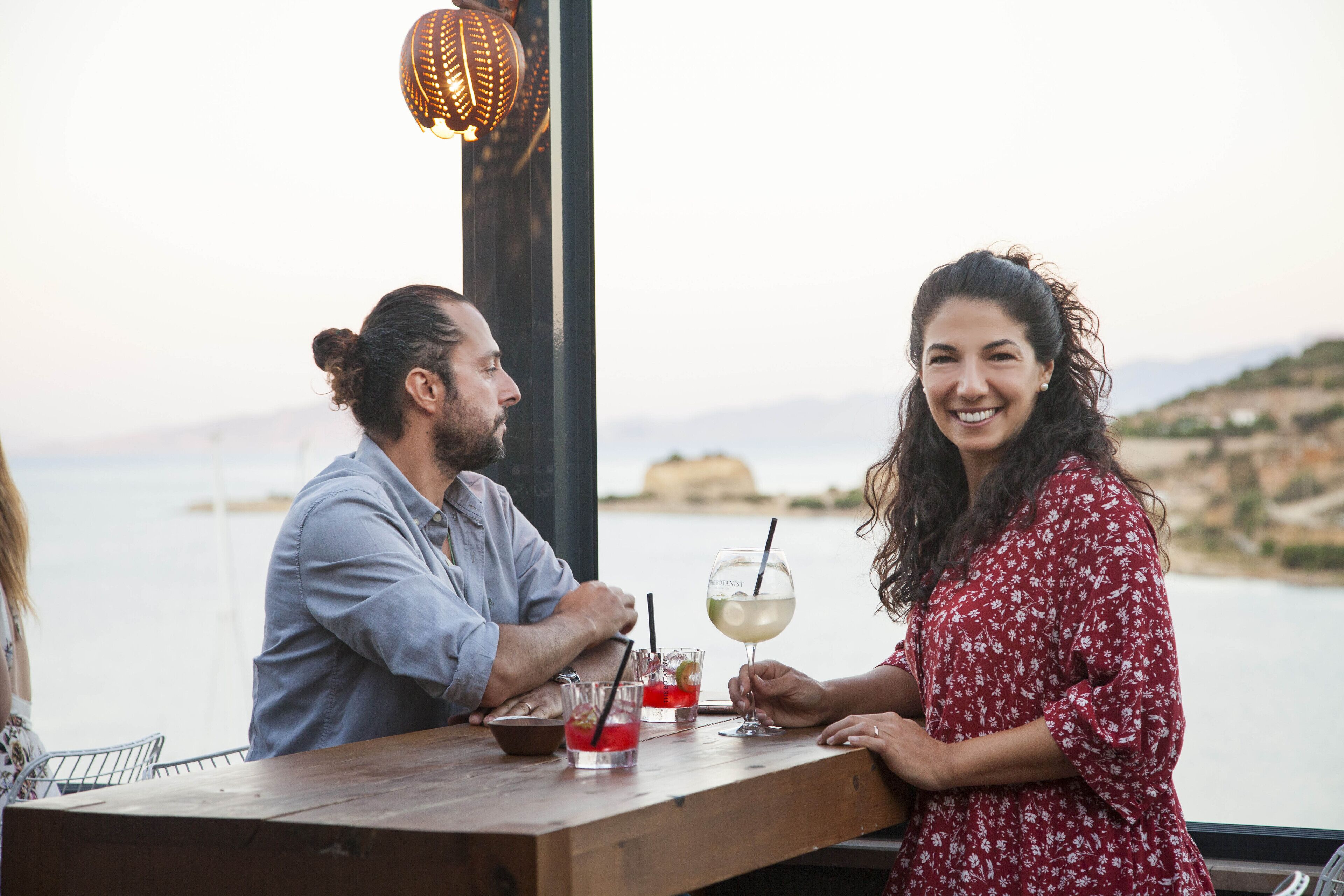 Una pareja sentada en una mesa de madera, con bebidas, mirando un cuerpo de agua y colinas.