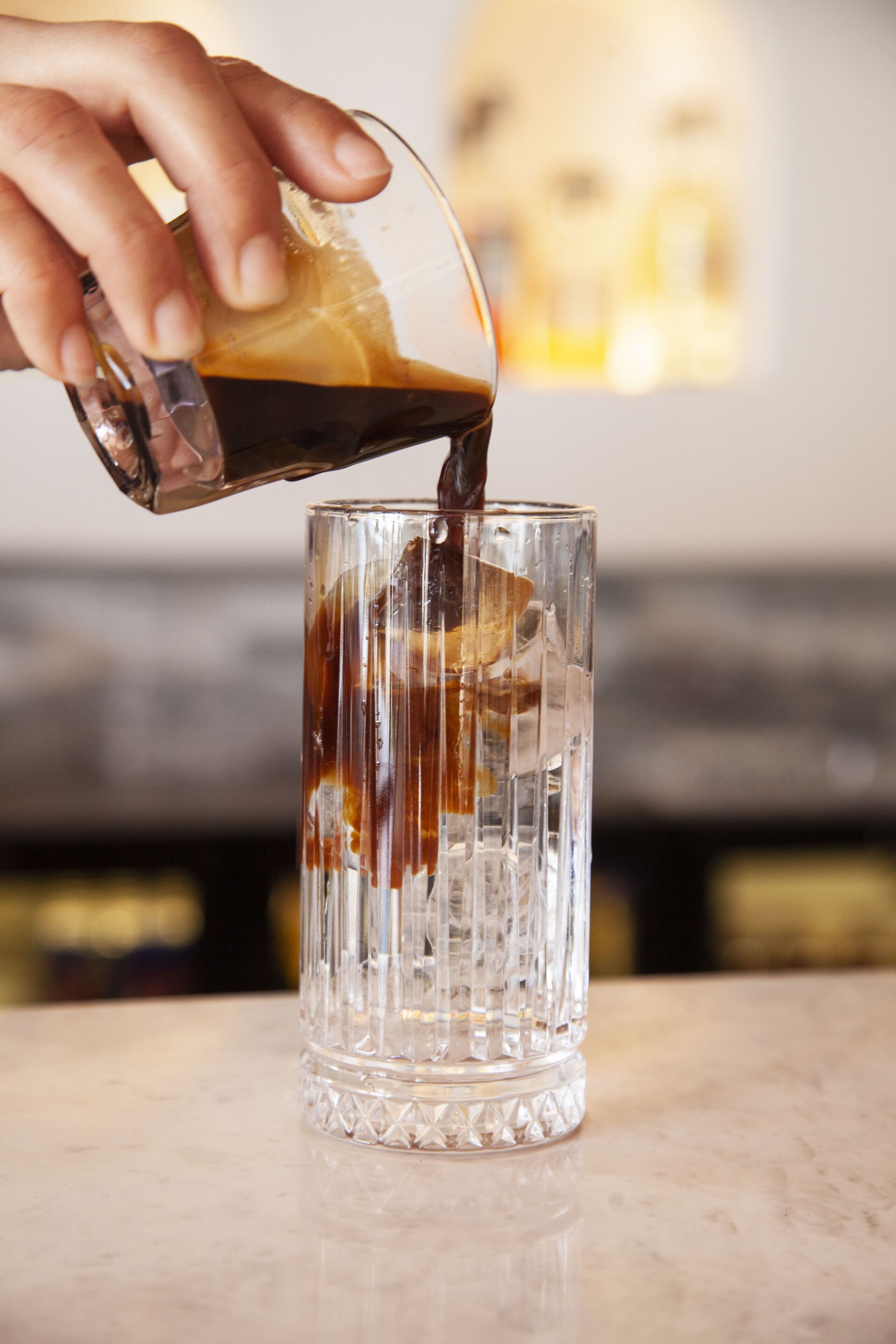 A hand pouring a dark liquid into a clear glass with a textured design.