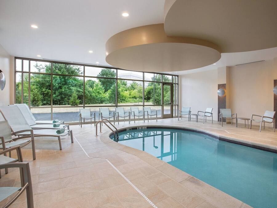 Piscina interior moderna com paredes de vidro transparente, cadeiras de descanso e vista para o verde lá fora.