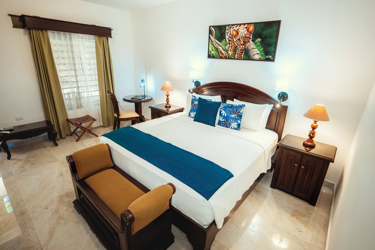 Une chambre d’hôtel avec un grand lit, deux tables de chevet avec des lampes, une chaise, un bureau et une fenêtre avec des rideaux.