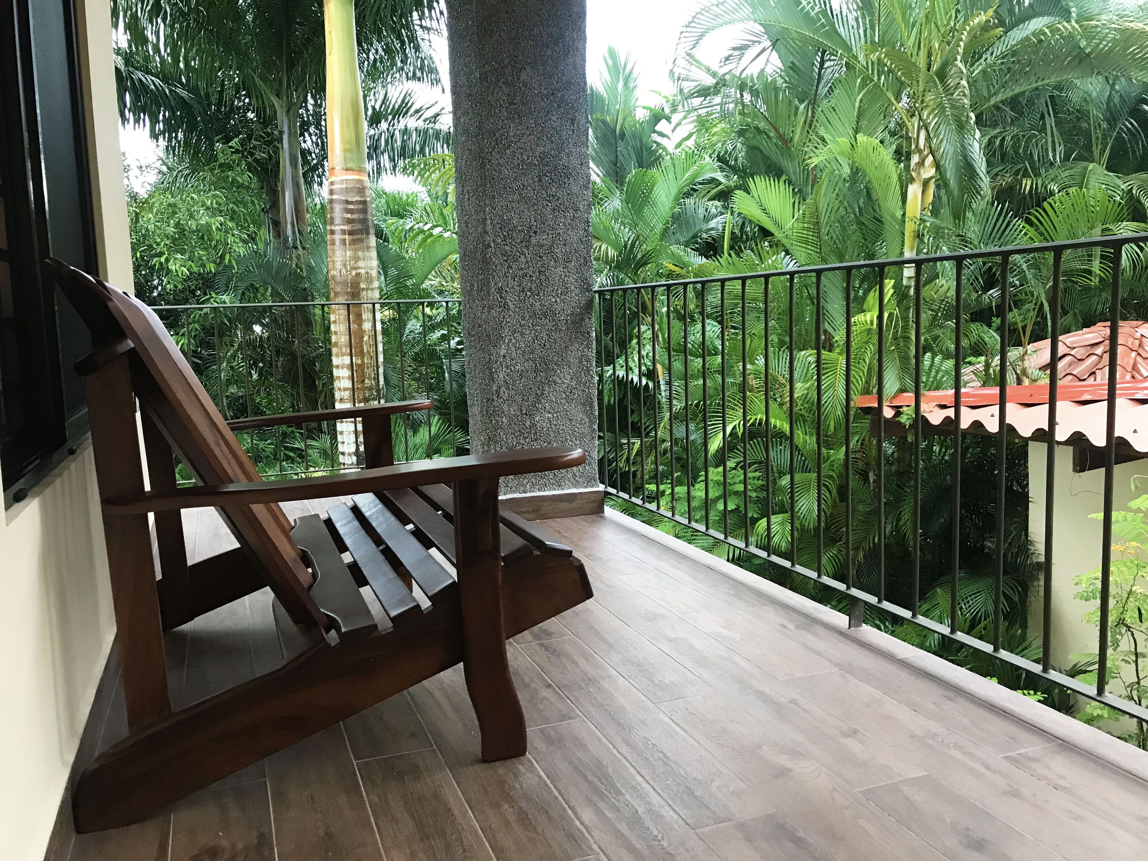 A wooden bench on a balcony with a view of lush greenery and a tiled floor.