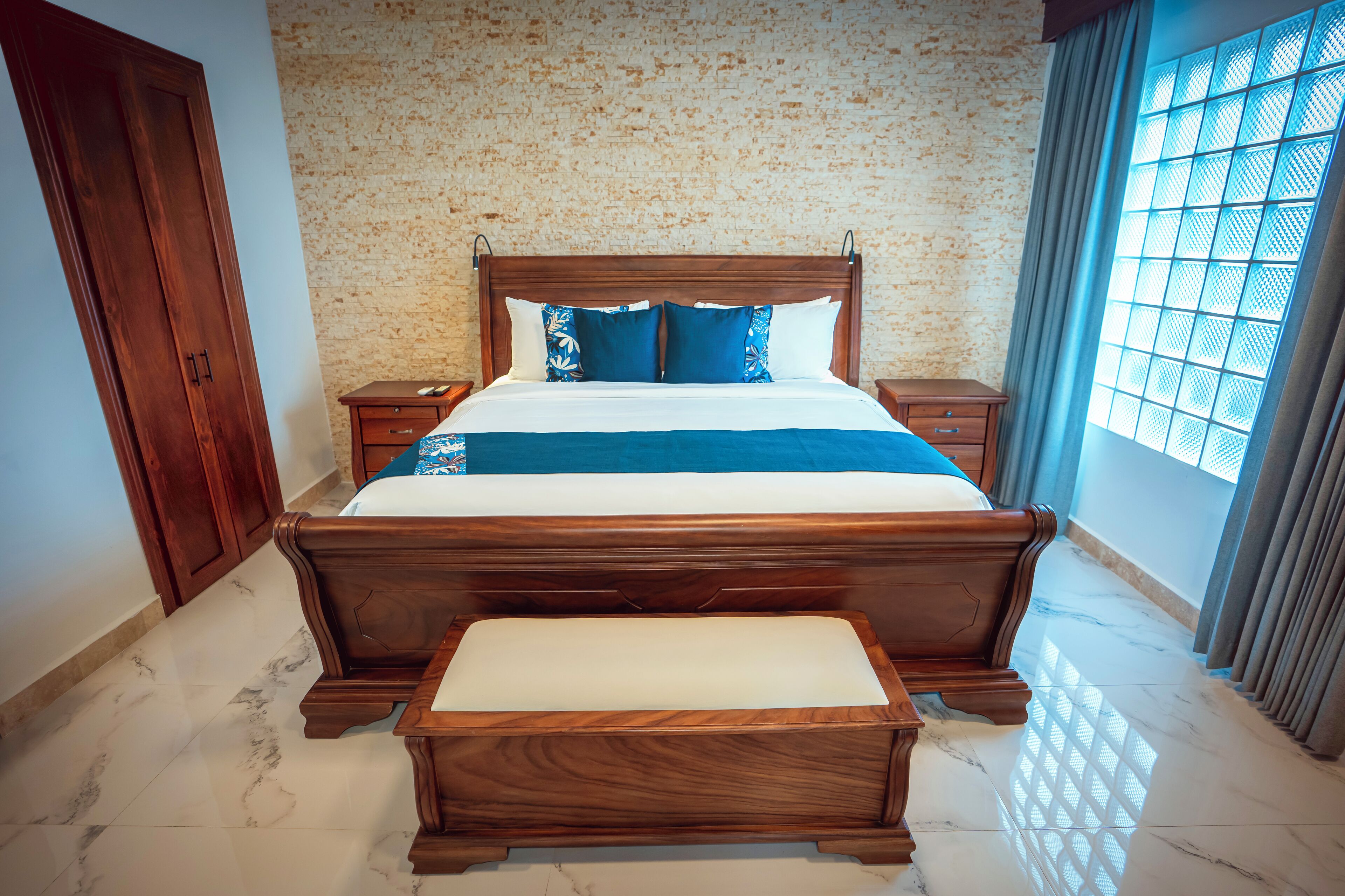 Un dormitorio con cama de madera, mesita de noche, banca, armario y ventana con cortinas.