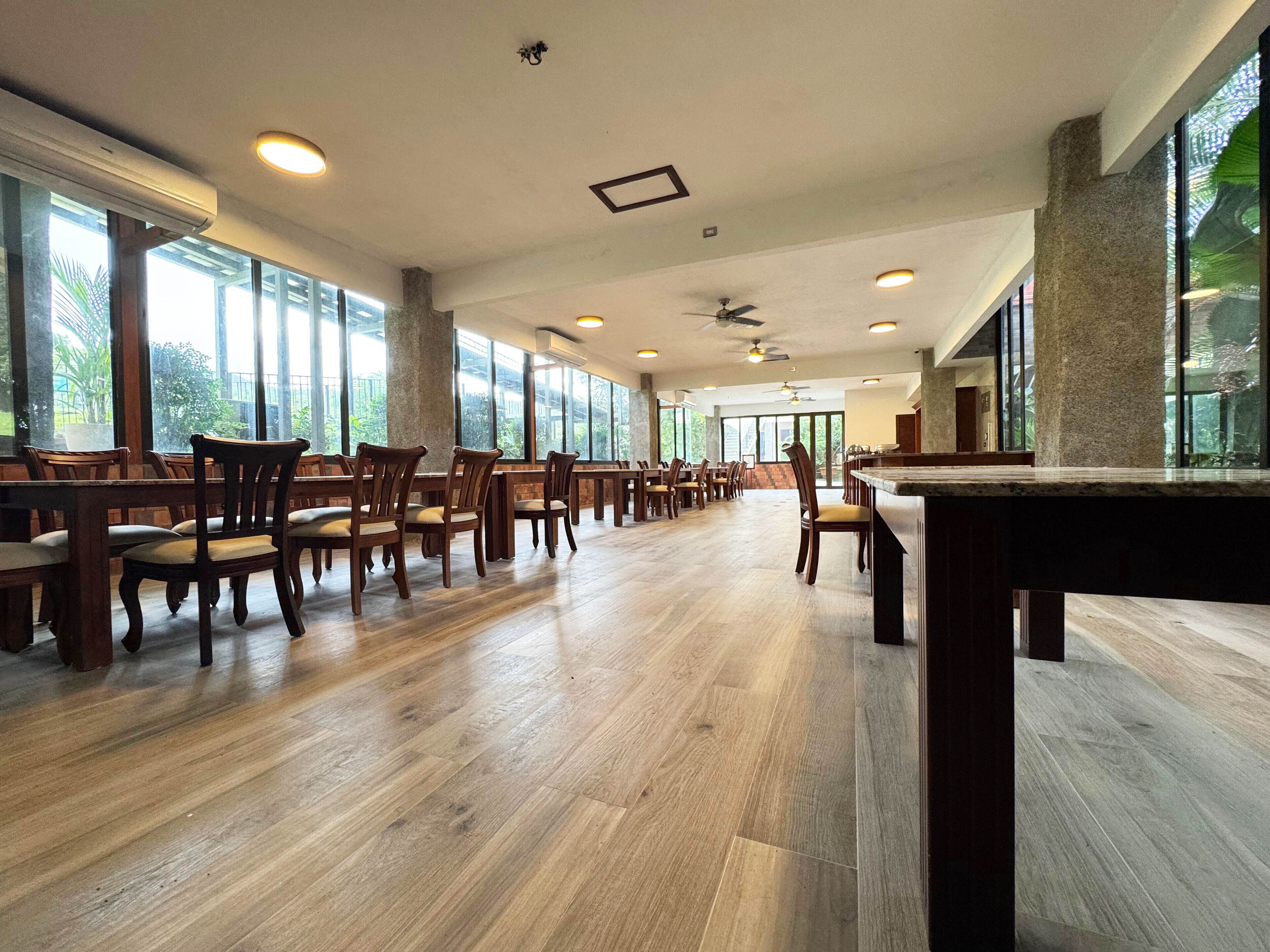 A spacious indoor area with wooden flooring, multiple tables and chairs, large windows, and ceiling fans.