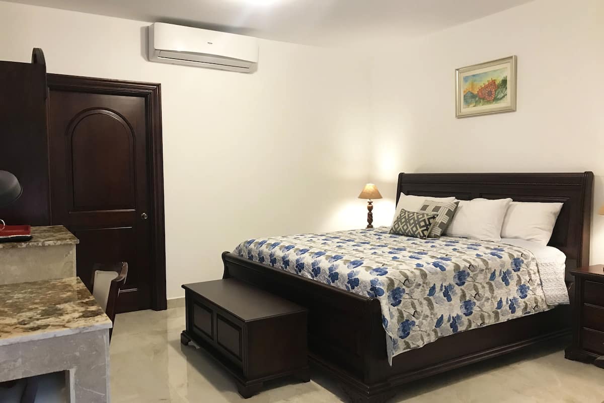Une chambre à coucher avec un grand lit, une table de chevet, une lampe, une chaise, une petite table, un climatiseur et un tableau accroché au mur.