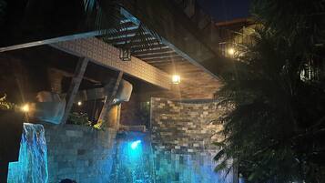 Zona de piscina exterior nocturna con una cascada, rodeada de muros de piedra y palmeras.
