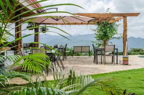 An outdoor patio with a wooden pergola, tables, chairs, and a view of mountains.