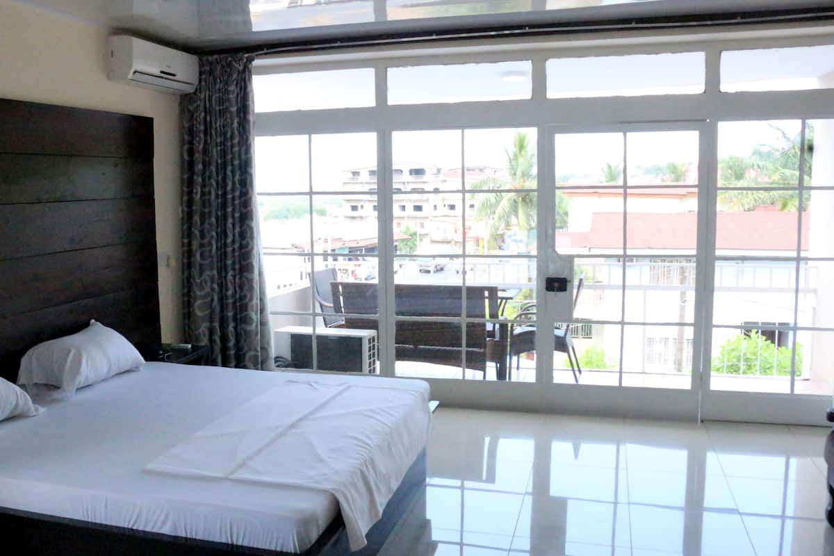 A hotel room with a large bed, a window with a view of buildings and greenery, and an air conditioning unit on the wall.