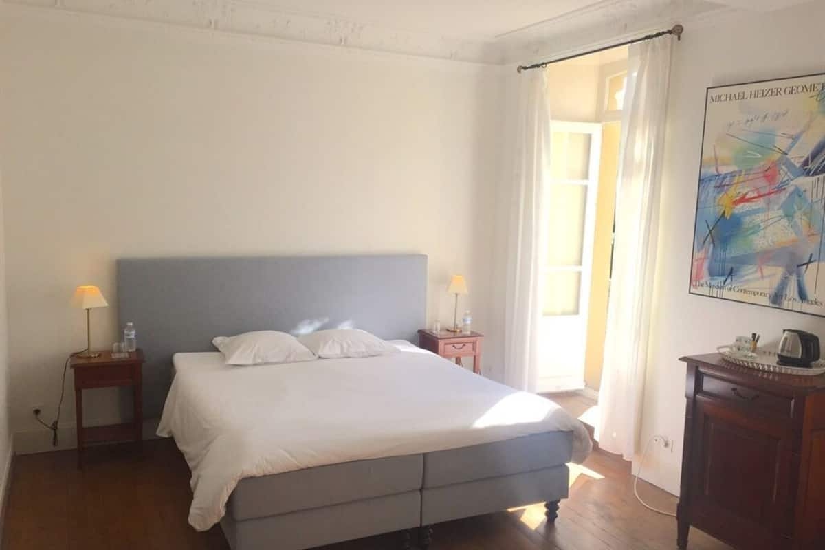 Een slaapkamer met een bed, nachtkastjes, een lamp, een poster aan de muur en een deur die naar een andere kamer leidt.