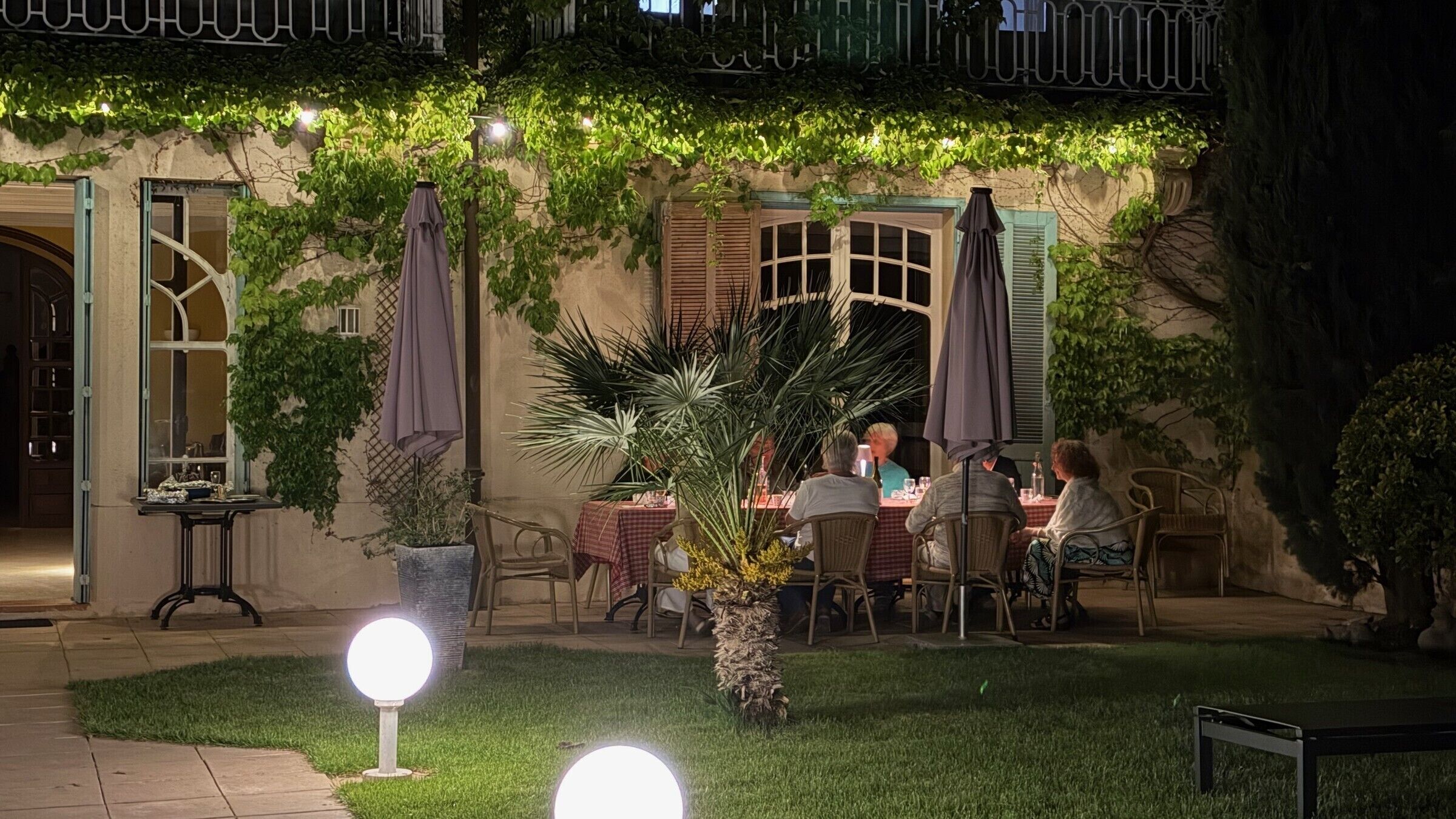 An outdoor dining area with tables and chairs, surrounded by greenery and a stone wall.