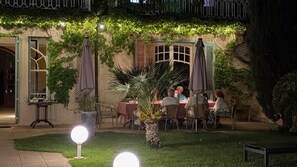 Un área de comedor al aire libre con mesas y sillas, rodeada de vegetación y un muro de piedra.