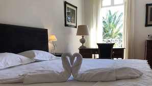 A hotel room with a neatly made bed, a desk with a lamp, and a view of palm trees through the window. - Le Jardin D'homps (Homps)