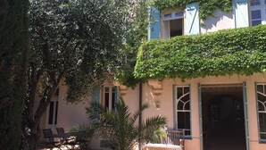 A building with a covered patio area, outdoor seating, and a large tree. - Le Jardin D'homps (Homps)