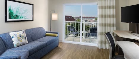 A hotel room with a blue sofa, a desk with a chair, a TV, a large window with a view of buildings, and a framed picture on the wall.
