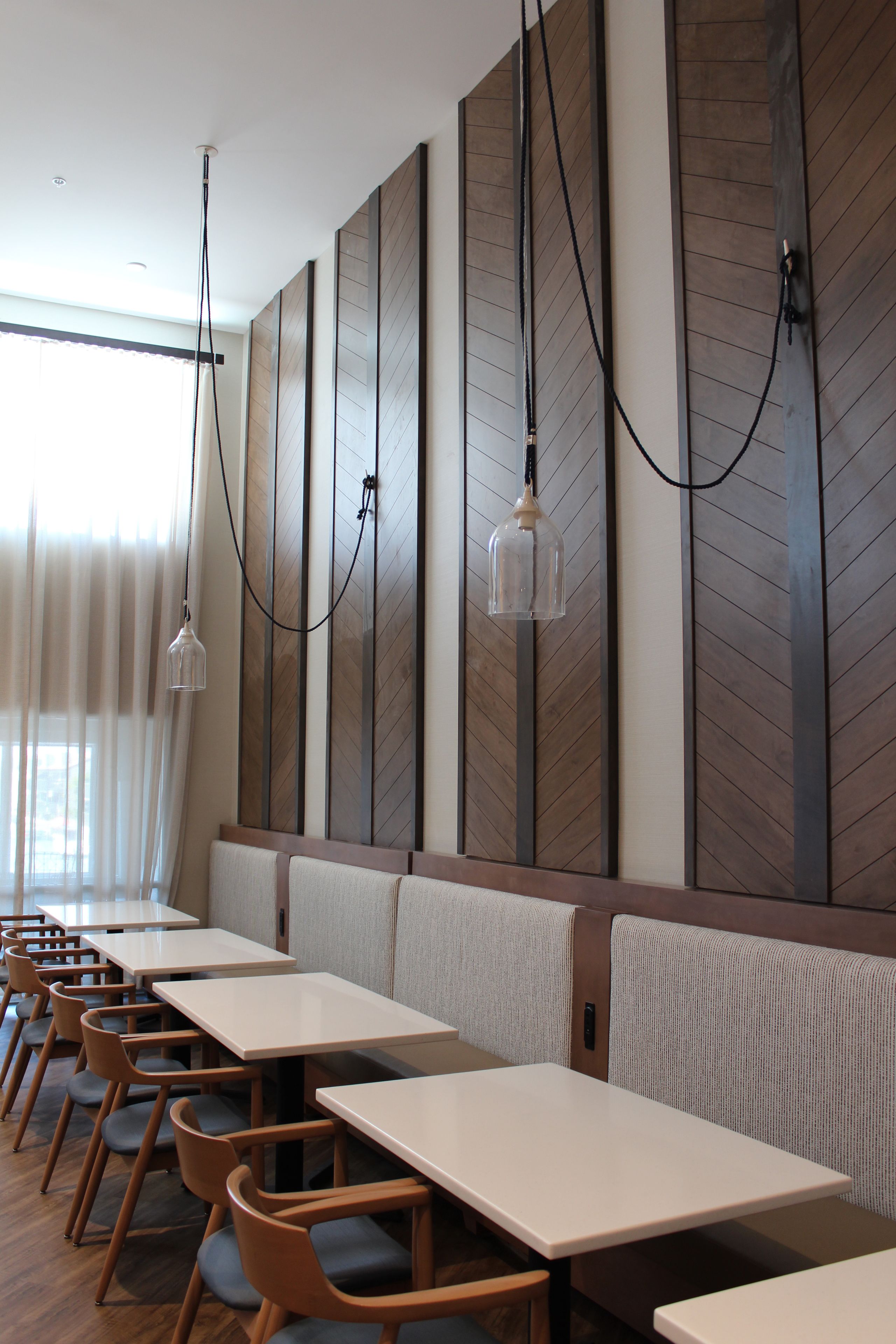 Et moderne restaurantinteriør med lange borde og siddepladser, vægpaneler i træ og hængende lamper.