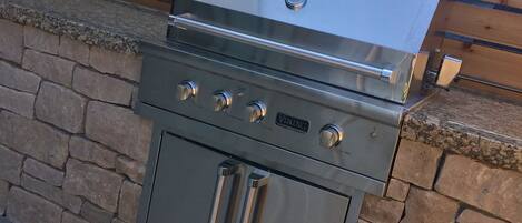 Una parrilla de acero inoxidable para exteriores con cuatro quemadores, un horno integrado y un quemador lateral, ubicada frente a un muro de piedra con una terraza de madera.