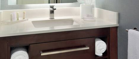 A modern bathroom with a large mirror, white sink, and dark wooden vanity. There are towels neatly rolled and stored in the vanity.