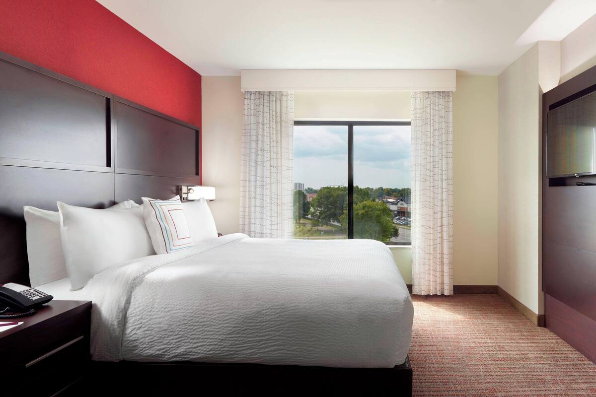 A hotel room with a large bed, a bedside telephone, a TV, and a window with curtains.