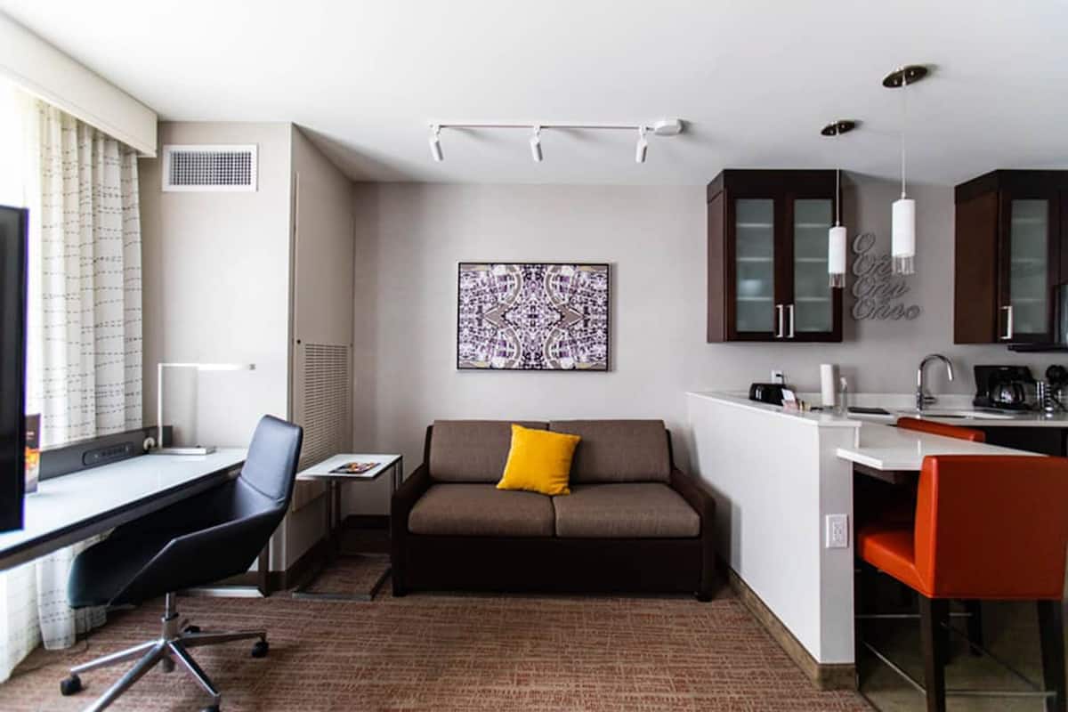 A compact hotel room with a desk, a sofa, a small kitchen area, and a large window with curtains.