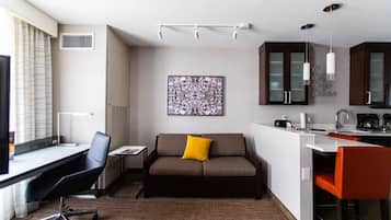 A compact hotel room with a desk, a sofa, a small kitchen area, and a large window with curtains.