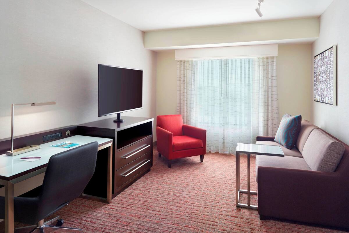A hotel room with a desk, a television, a red armchair, a sofa, and a window with curtains.