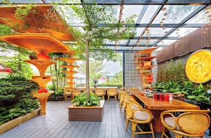 Un espacio exterior para comer con mobiliario de mimbre, una jardinera central con follaje y una estructura decorativa colgante.