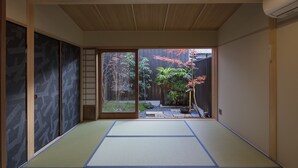 A traditional Japanese room with tatami flooring, sliding doors, and a view of a garden with plants and a wooden fence. - Shimabara Kaiden (Kyoto)