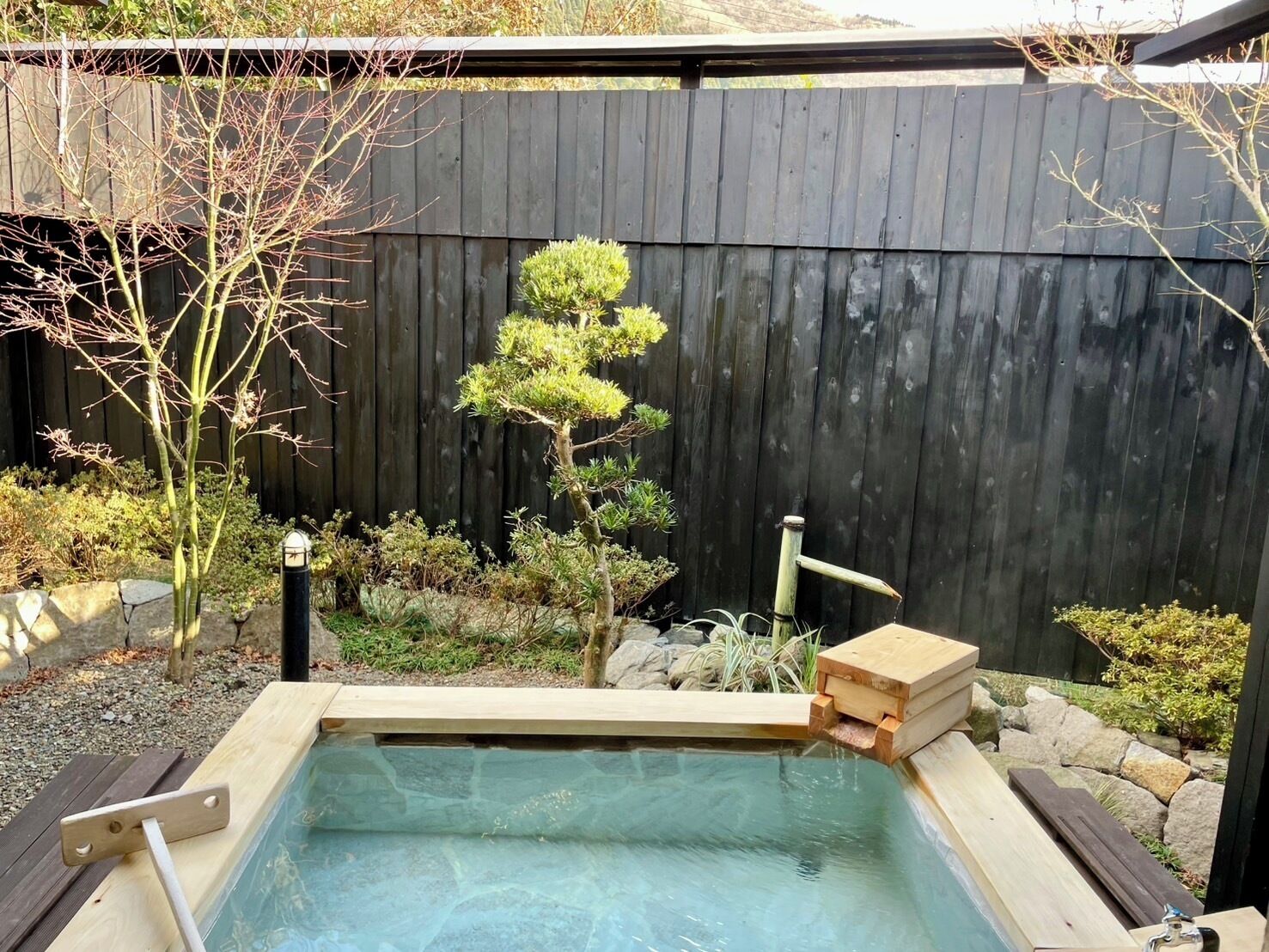 Un jacuzzi con una cubierta de madera, rodeado por una valla de madera, un árbol pequeño y varias plantas.