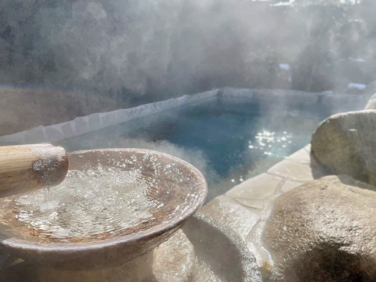 Um banho de águas termais com uma concha de madeira, vapor subindo da água e ladrilhos de pedra ao redor.