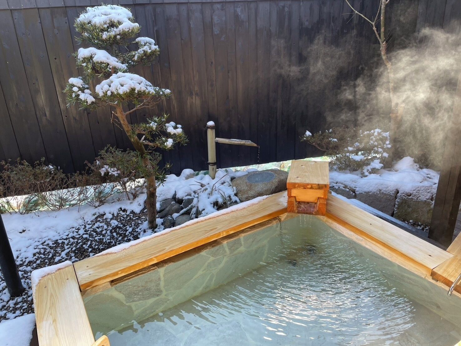 Un espace de bains en eau chaude, doté d’une structure en bois, entouré d’un sol enneigé et d’une clôture en bois.