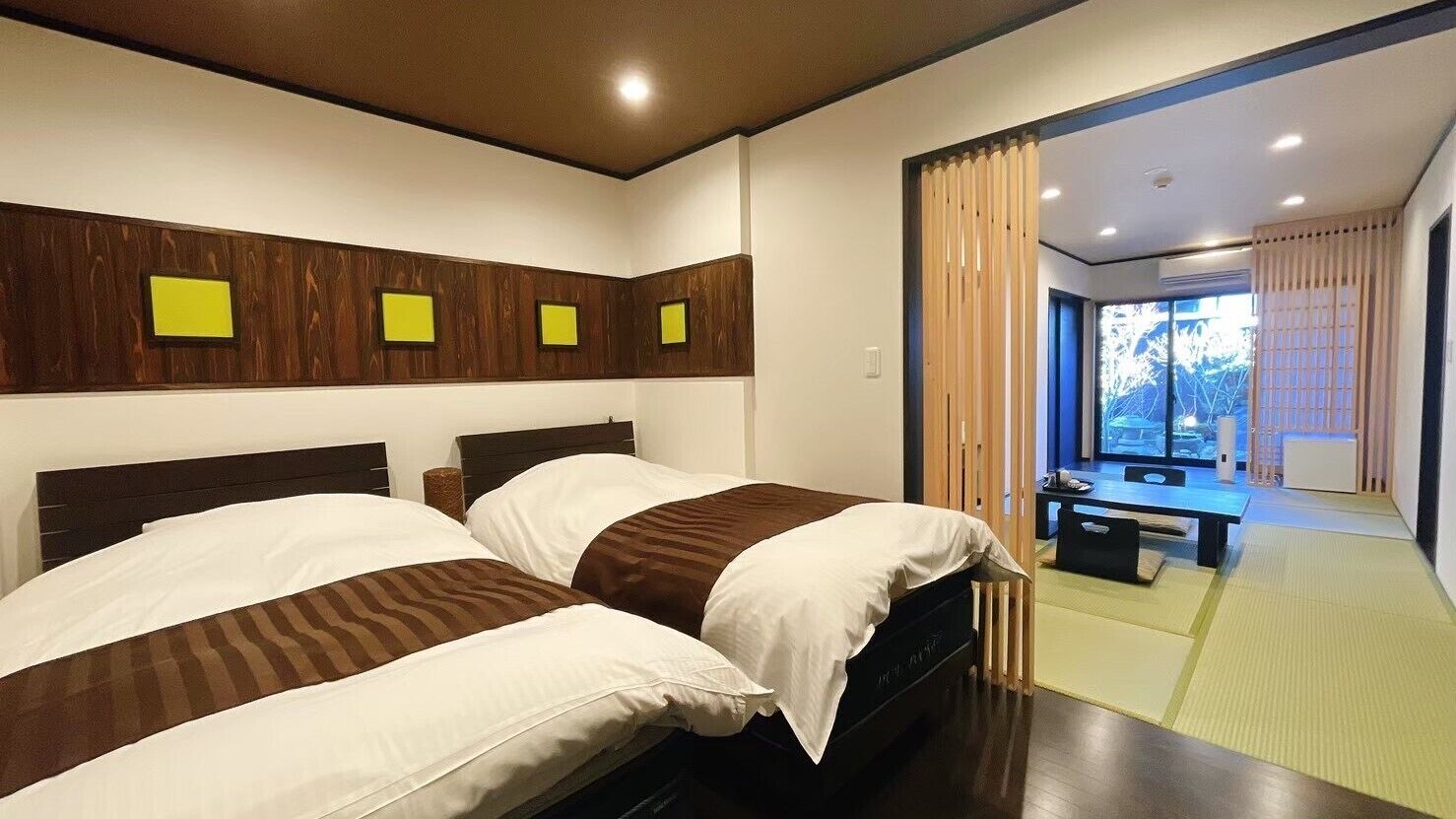 A hotel room with two beds, a wooden headboard, a small table, and a view through a window.