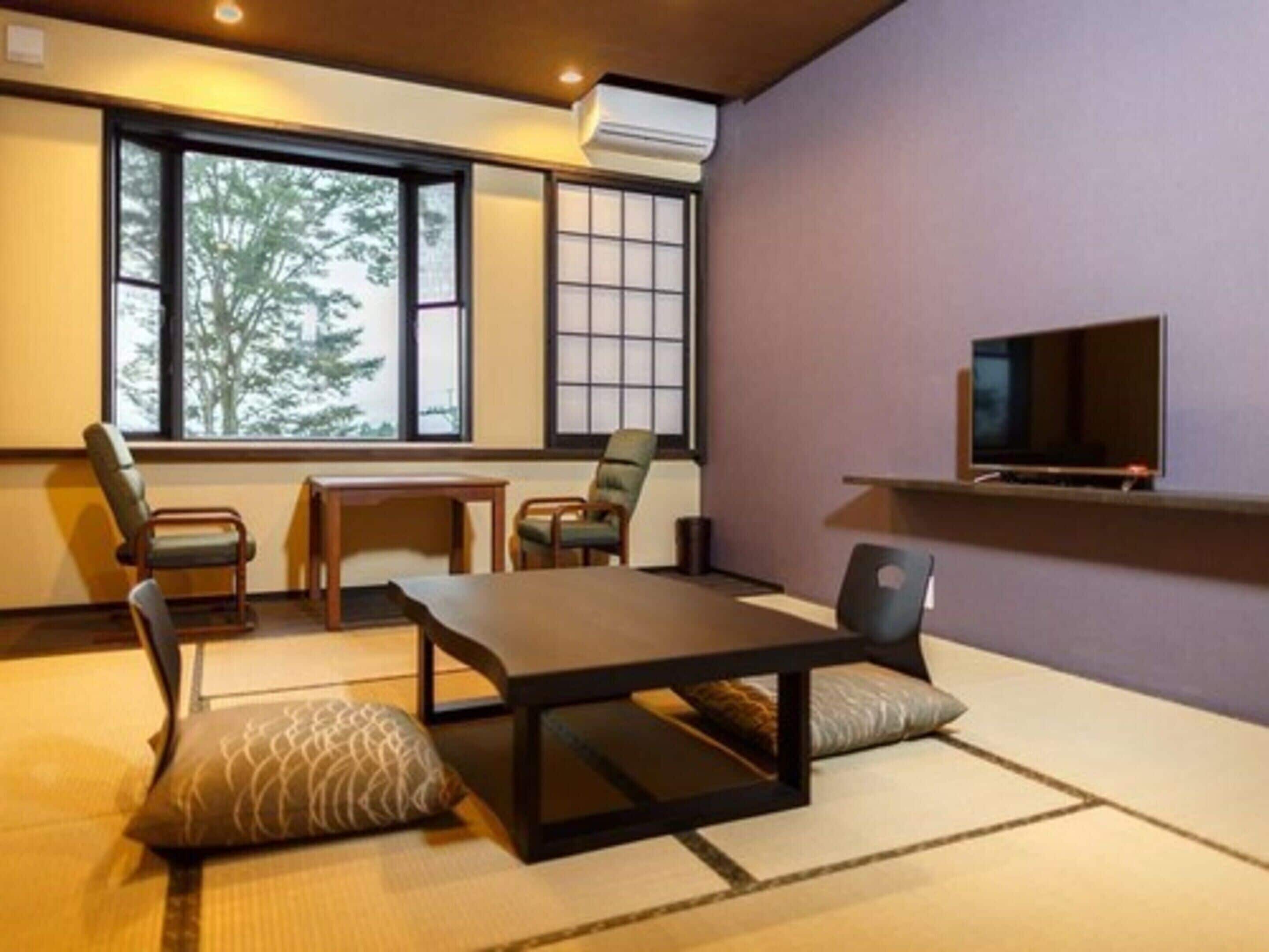 A room with tatami mats, a low table, chairs, a television on a shelf, and a window with a view of trees.