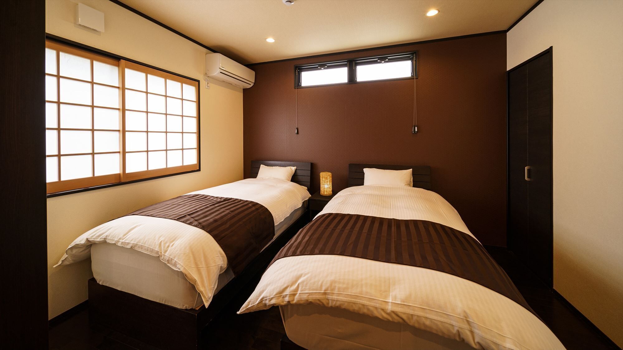 A hotel room with two beds, a window, an air conditioning unit, and a door.