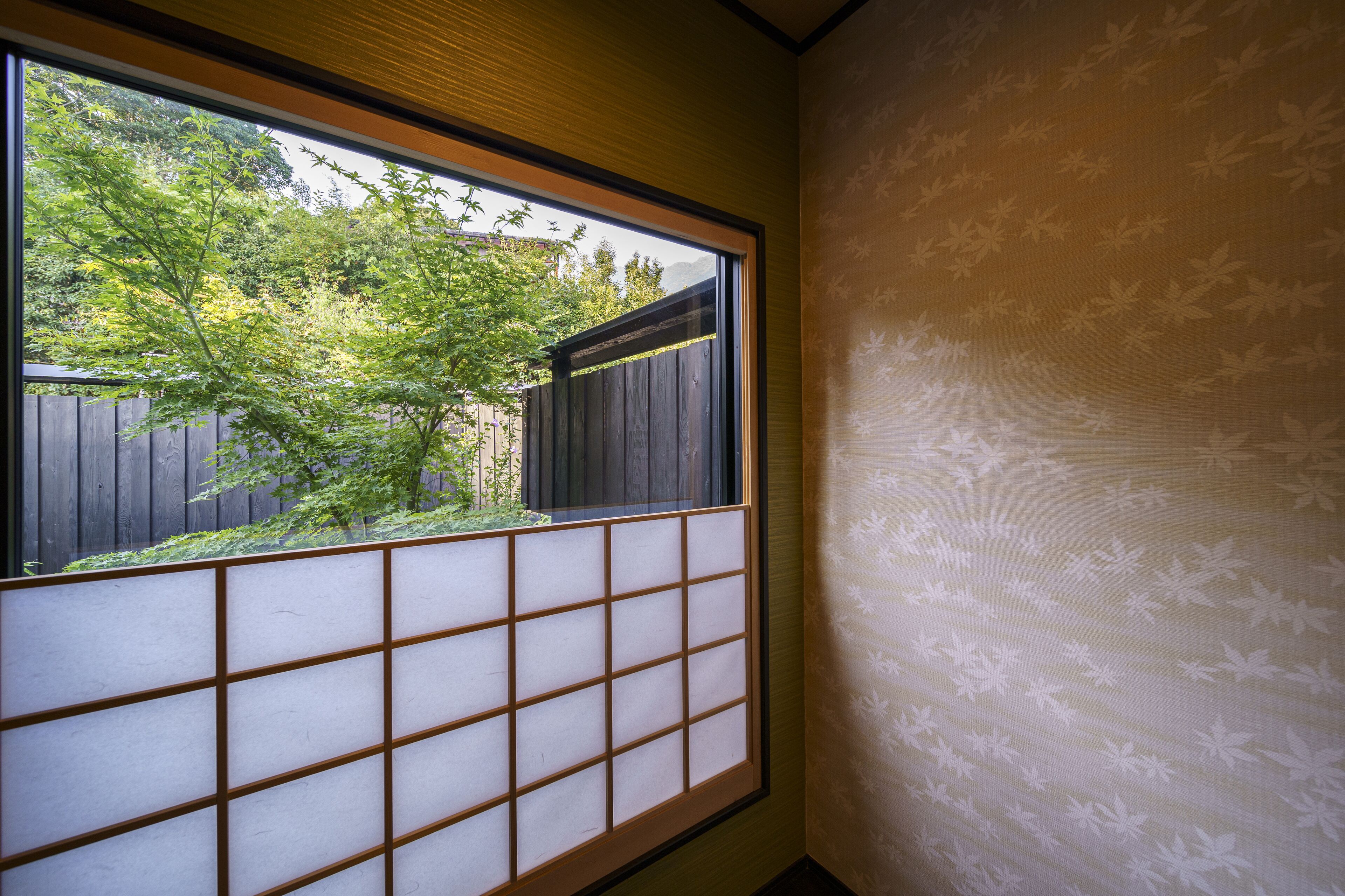 Une pièce dotée d’une fenêtre à panneaux coulissants de style japonais, d’une clôture en bois à l’extérieur et d’un mur orné d’un motif discret représentant des feuilles.