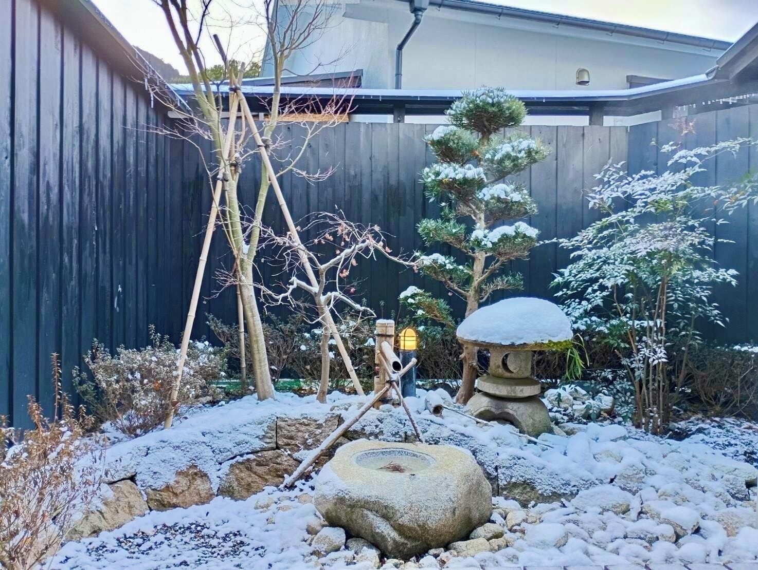 Un jardín cubierto de nieve con un farol de piedra, un árbol bonsái y una valla de madera.