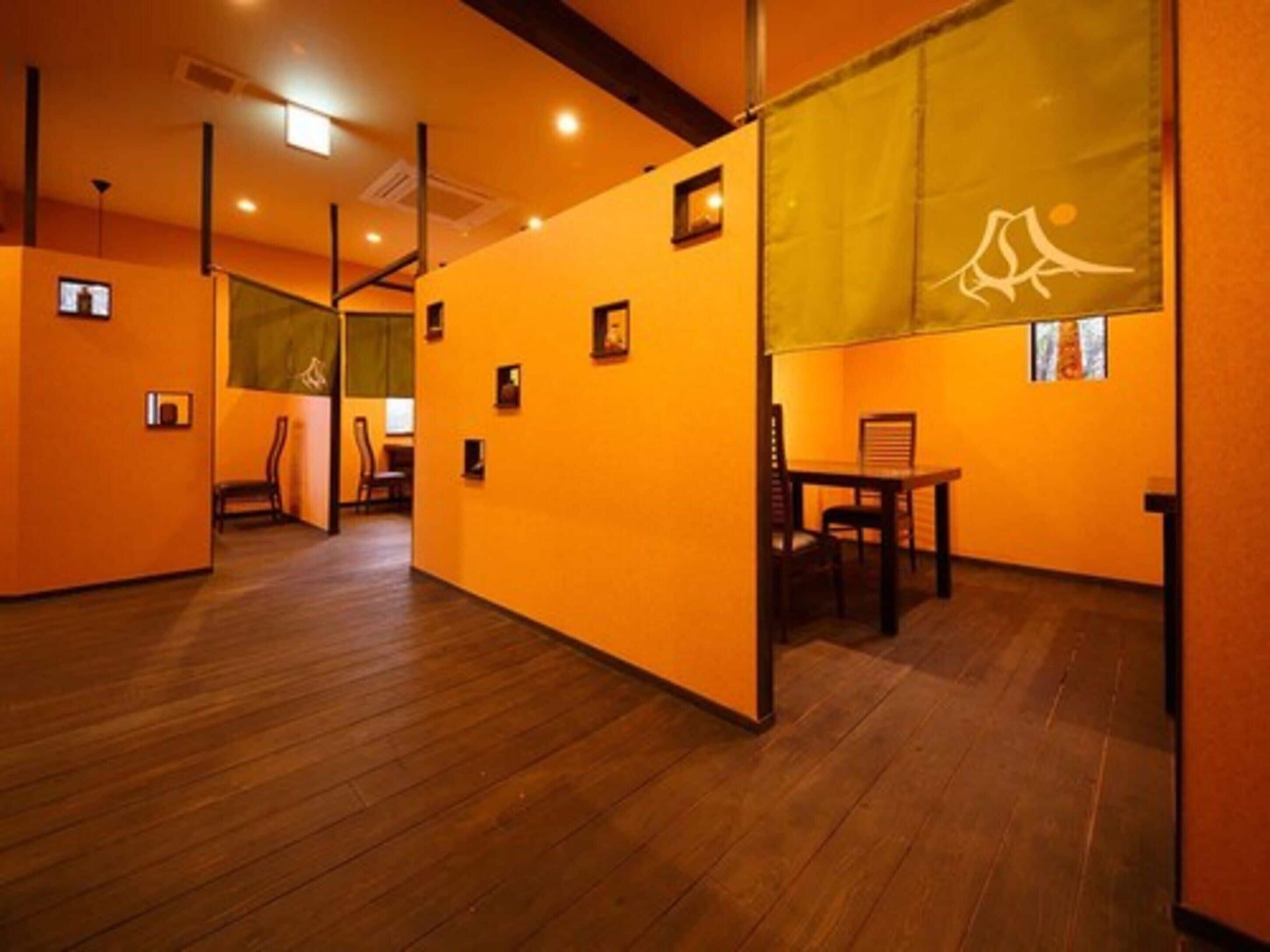 A room with wooden flooring, orange walls, and a large window with a curtain. There are framed pictures on the walls and a chair in the corner.