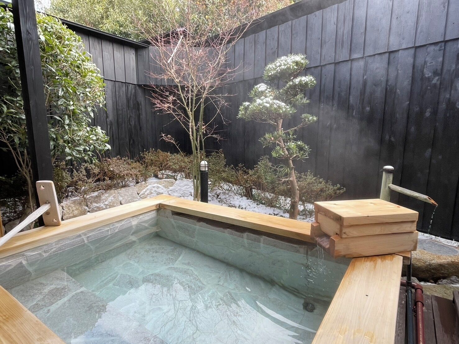 A hot tub with a wooden deck, surrounded by a wooden fence and various plants.