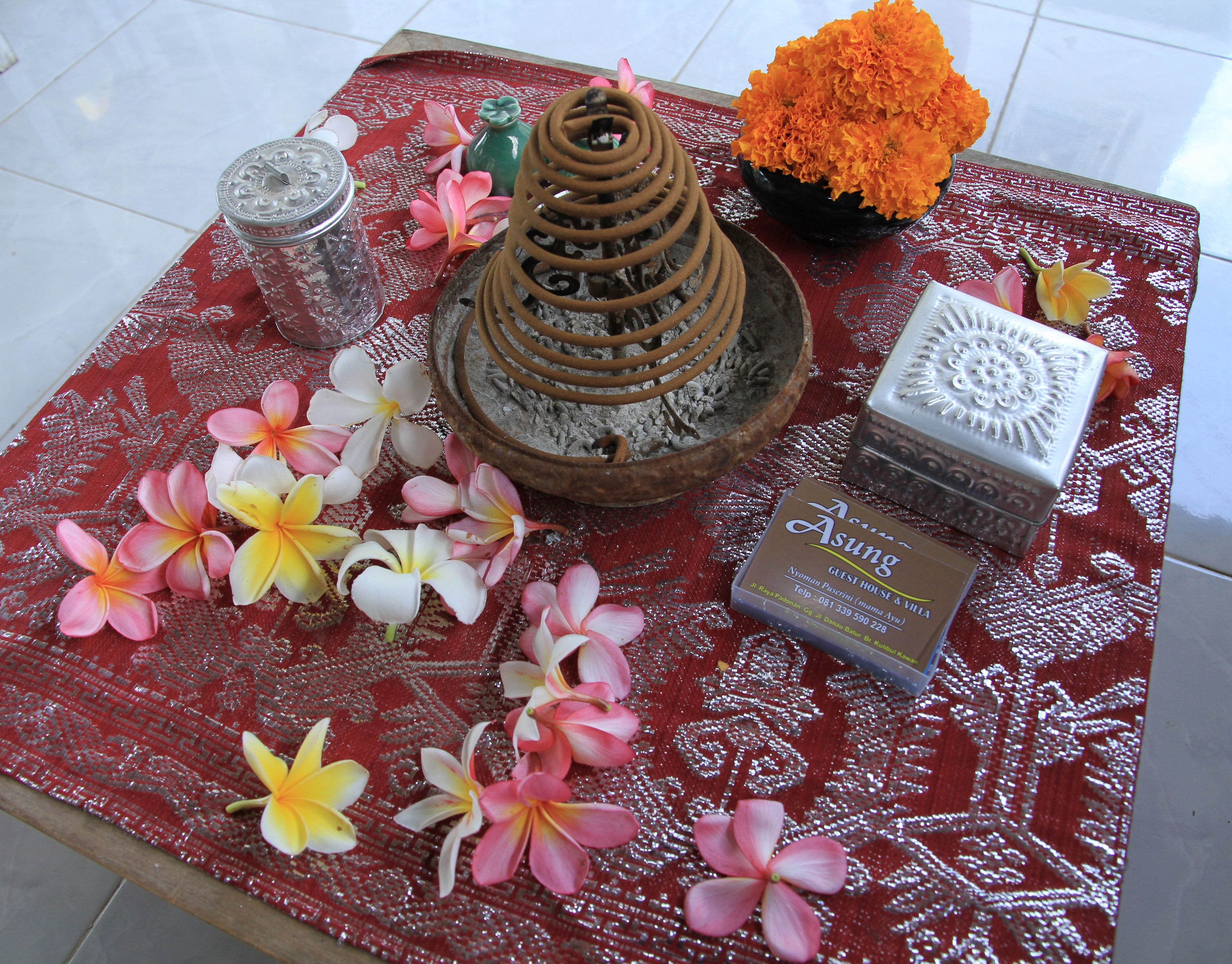 A table with a red patterned tablecloth, a decorative incense burner, a bowl of orange flowers, a small green vase, and a card with hotel information.