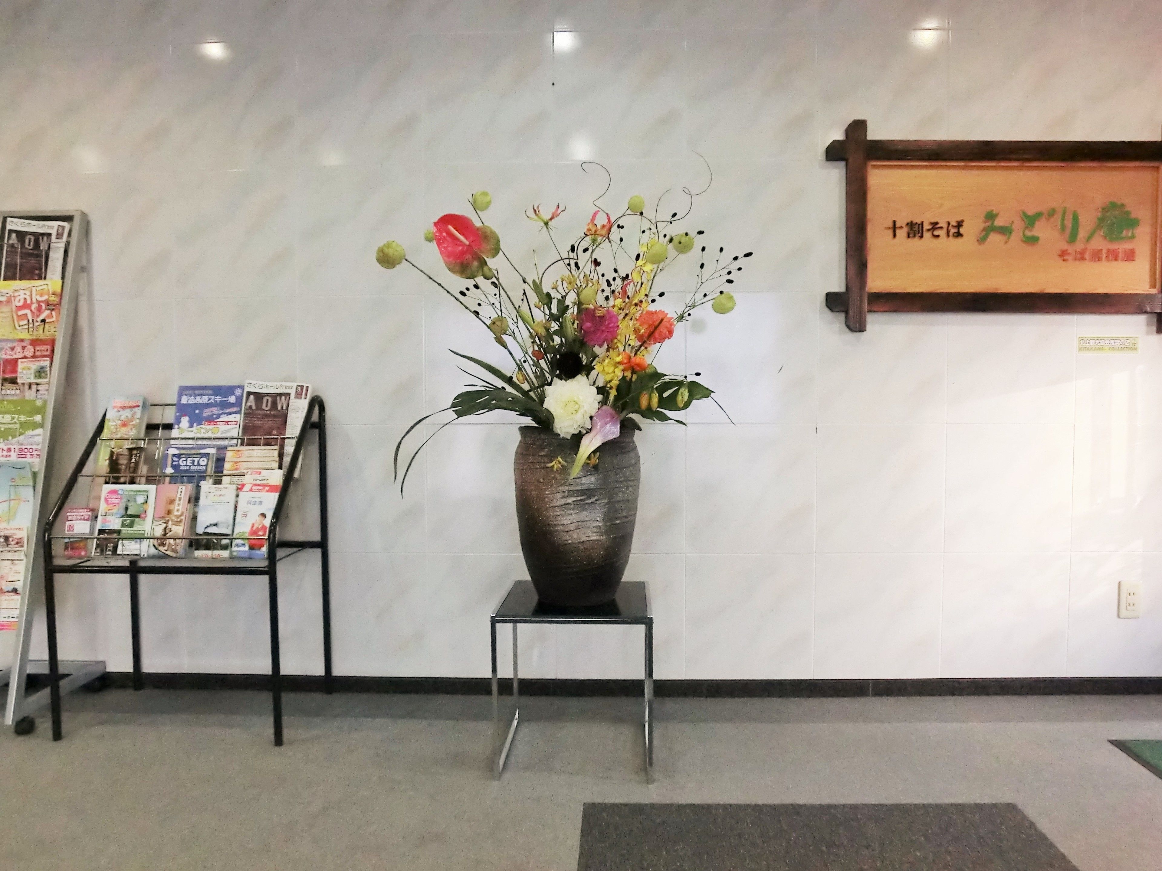 Un área de recepción con un jarrón de flores sobre un pedestal, un atril para revistas y un letrero con caracteres japoneses.