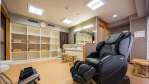 A massage therapy room with a massage chair, shelves with baskets, a small table, and a fan.