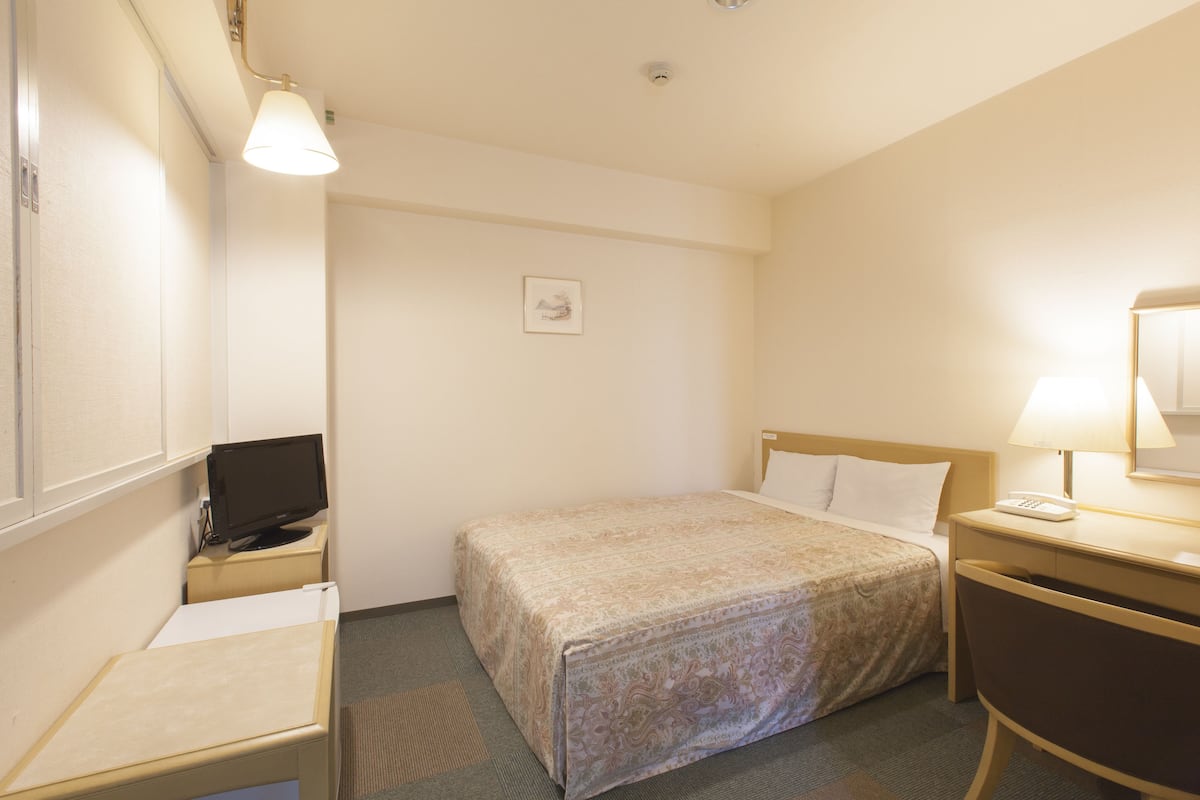 A hotel room with a bed, bedside table, lamp, and a desk with a computer.