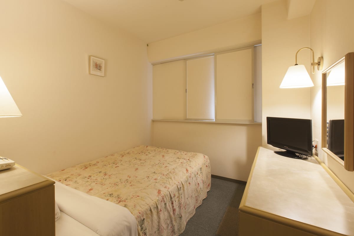 A small, neatly arranged hotel room with a single bed, a bedside table with a lamp, a television, and a window with blinds.