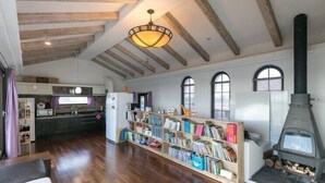 Ein geräumiger Raum mit Holzbalken an der Decke, einem großen Bücherregal, einem Kamin und einer Küchenzeile mit Kühlschrank und Spüle.