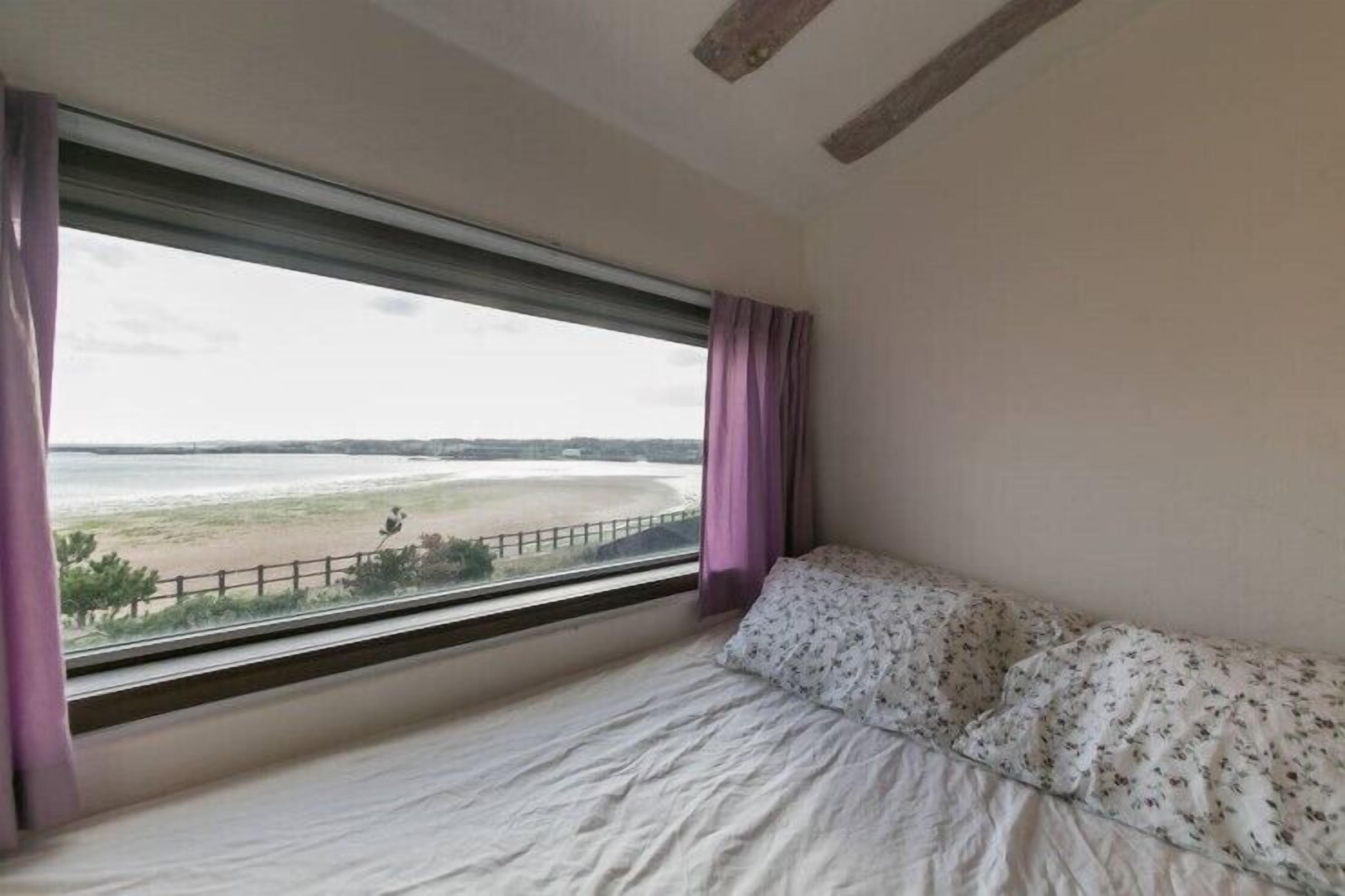 A bedroom with a bed, a window with a view of a beach, and a wooden ceiling beam.