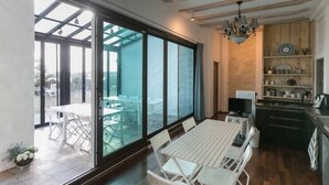 Ein geräumiges Zimmer mit einer großen Schiebeglasdoor, die auf eine Terrasse im Freien führt. Die Holzbalkendecke beherbergt eine Kronleuchter. Rechts ist eine Küchenzeile mit Holzschränken und einer Arbeitsplatte erkennbar.