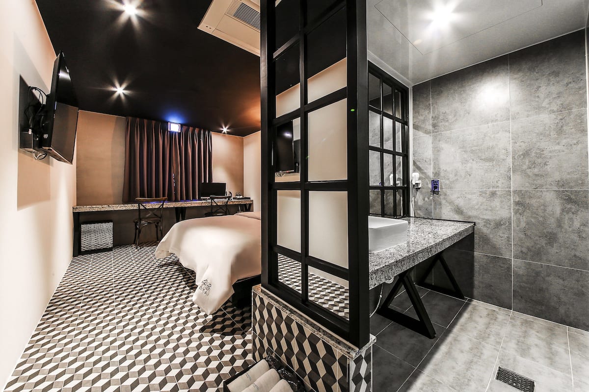 A modern hotel room with a checkered floor, a bed with a white cover, a desk with a computer, and a bathroom with a granite countertop and tiled walls.