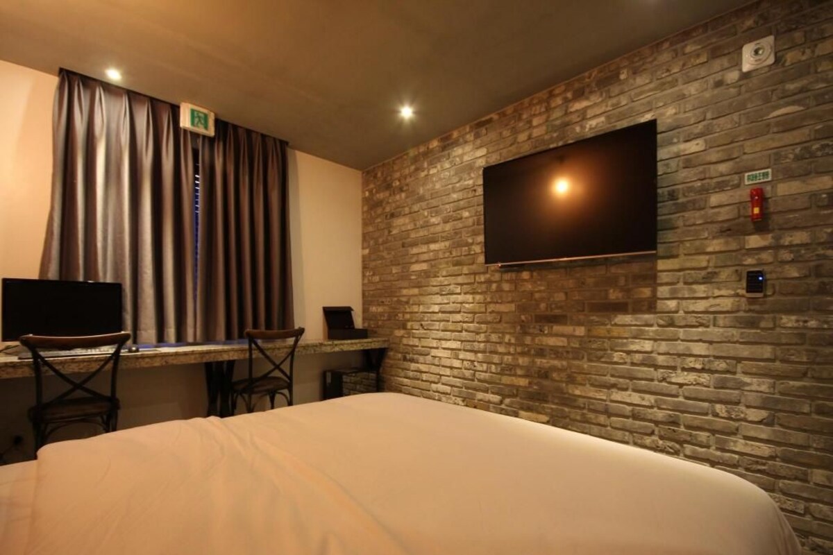 A hotel room with a large bed, a desk with a computer, and a flat-screen TV mounted on a brick wall.